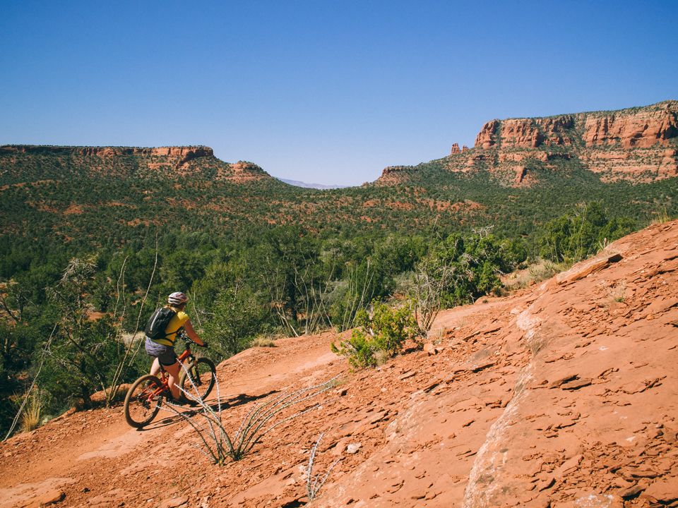 Cyclist in a yellow shirt rides a red mountain bike down a red dirt desert trail toward green mesas under a clear blue sky