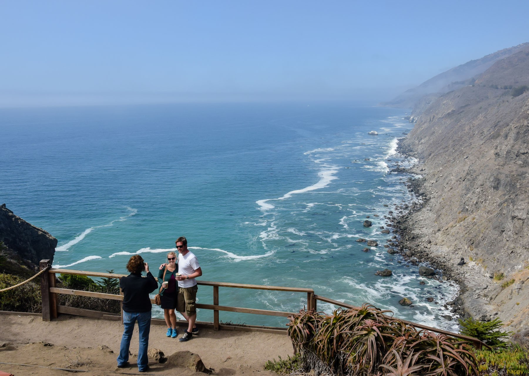 Three people at a wooden fence viewpoint as one photographs a couple overlooking turquoise ocean waves and rocky coastal cliffs